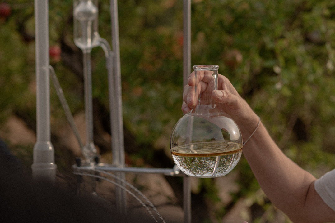 A hand holding a glass flask at the distillation still, outdoors at Ortofficina