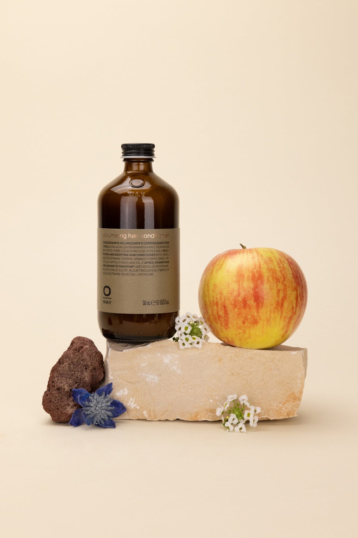 Volumizing hair conditioner in an amber glass bottle on a block of stone, beside an apple, a cornflower and small white flowers.